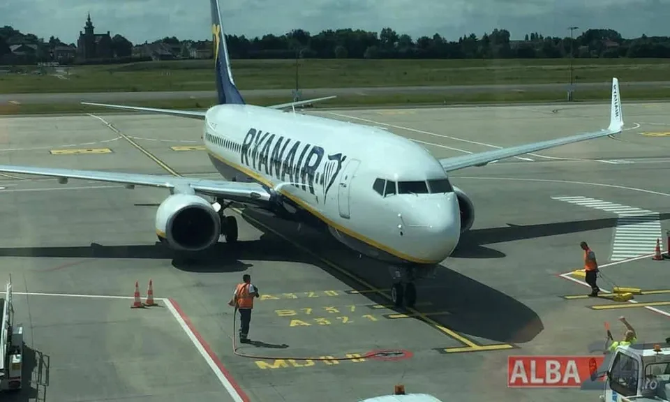 Un avion alb cu inscripția Ryanair stă pe pista unui aeroport lângă un angajat care poartă o vestă portocalie.