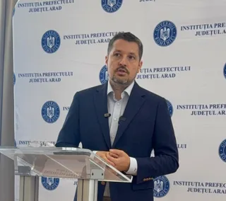 Un bărbat cu sacou închis la culoare și cămașă deschisă stă în spatele unui pupitru transparent în fața unui panou cu sigilii oficiale.