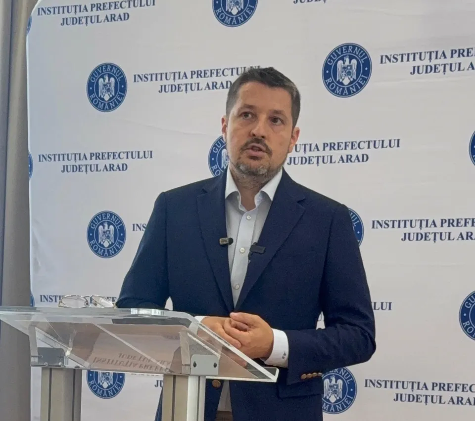 Un bărbat cu sacou închis la culoare și cămașă deschisă stă în spatele unui pupitru transparent în fața unui panou cu sigilii oficiale.