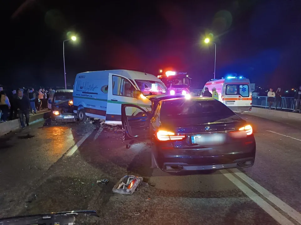 Un autoturism negru și un furgon alb sunt poziționate pe o stradă noaptea, lângă o ambulanță cu luminile aprinse.