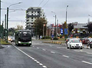 Un autobuz verde și negru circulă pe un drum asfaltat cu mașini albe și colorate în partea dreaptă, sub un cer înnorat.