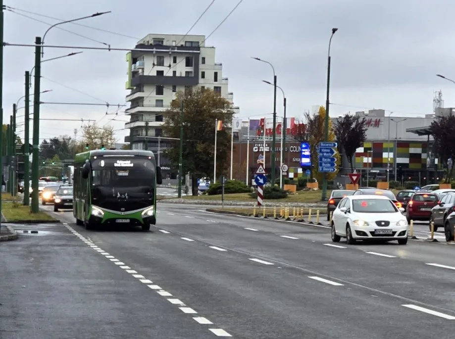 Un autobuz verde și negru circulă pe un drum asfaltat cu mașini albe și colorate în partea dreaptă, sub un cer înnorat.