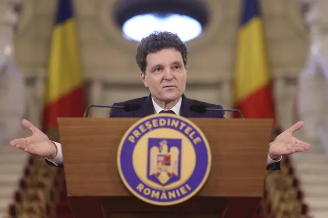 Un bărbat cu părul închis la culoare stă în spatele unui podium de lemn cu un emblema circulară și își întinde ambele mâini în lateral.