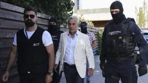Un bărbat cu părul alb și sacou deschis merge printre patru bărbați îmbrăcați în haine negre, dintre care doi poartă măști complete pe față.