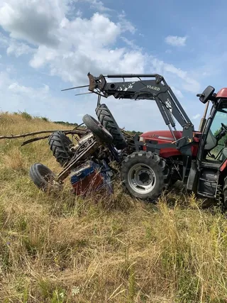 Un tractor roșu cu un braț hidraulic este răsturnat pe o laterală într-un câmp cu iarbă uscată sub un cer cu nori.