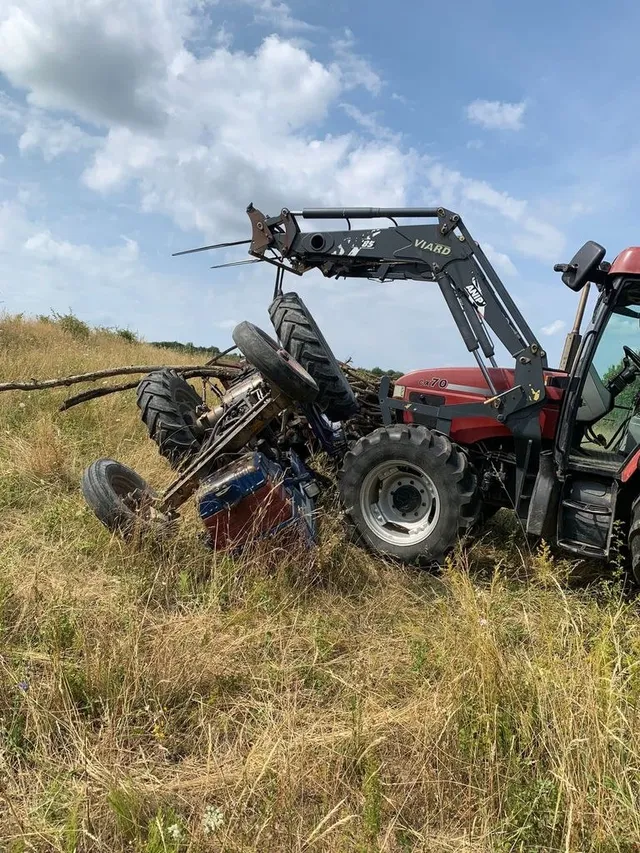 Un tractor roșu cu un braț hidraulic este răsturnat pe o laterală într-un câmp cu iarbă uscată sub un cer cu nori.