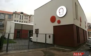 O clădire albă cu elemente circulare roșii și un logo rotund este situată în spatele unui gard metalic gri.