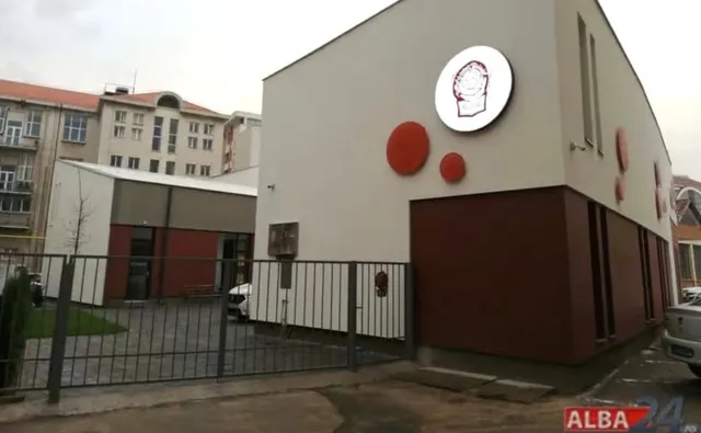 O clădire albă cu elemente circulare roșii și un logo rotund este situată în spatele unui gard metalic gri.