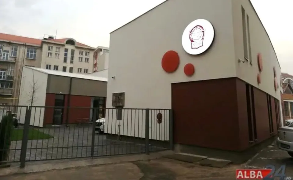 O clădire albă cu elemente circulare roșii și un logo rotund este situată în spatele unui gard metalic gri.