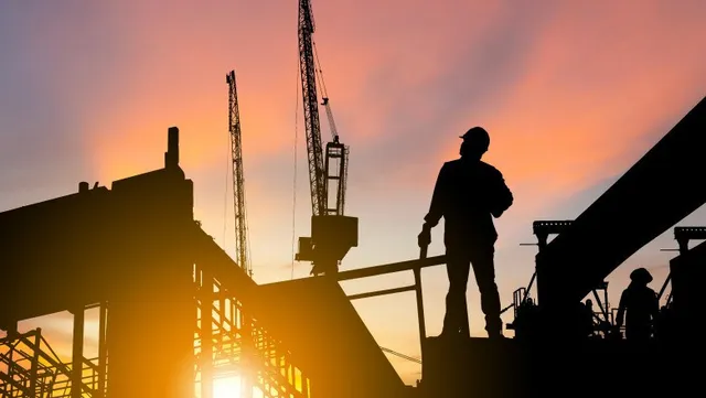 Siluetele unor lucrători și ale unor macarale de construcții se ridică în fața unui cer colorat la apus.