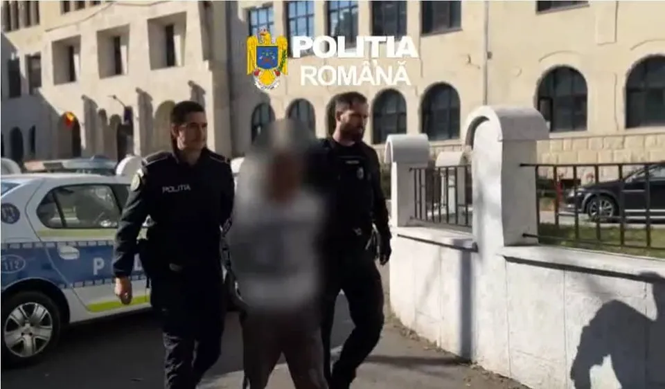 Doi polițiști în uniformă întunecată conduc un bărbat cu fața neclară pe un trotuar lângă un autovehicul alb și un gard de piatră.