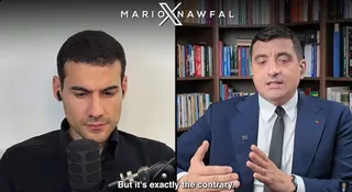 Un bărbat cu cămașă neagră privește în jos lângă un microfon, în timp ce un alt bărbat în costum și cravată vorbește în fața unei biblioteci.