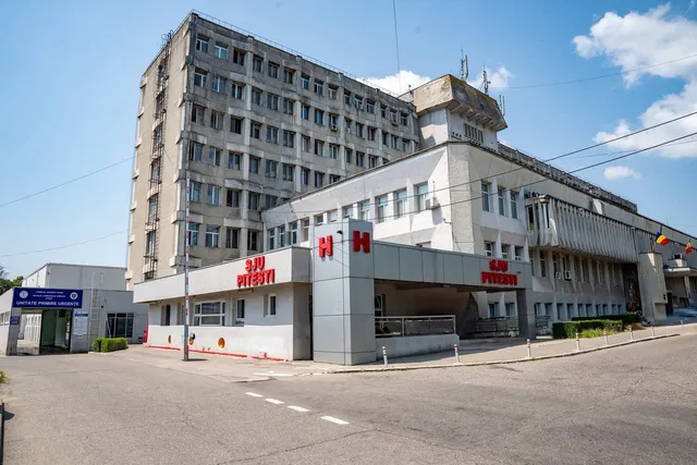O clădire înaltă de beton cu multe ferestre este situată lângă o structură mai joasă care prezintă un panou gri cu litere roșii și albe.
