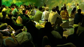 Oamenii stau pe saci de rahat și pe podea, privind o proiecție luminoasă cu vegetație pe un perete într-o încăpere întunecată.