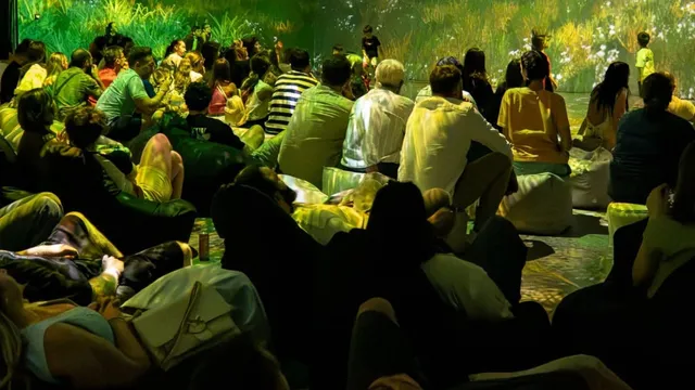 Oamenii stau pe saci de rahat și pe podea, privind o proiecție luminoasă cu vegetație pe un perete într-o încăpere întunecată.