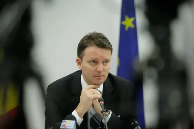 Un bărbat cu părul scurt, îmbrăcat în costum negru și cămașă albă, stă la o masă cu mâinile sub bărbie în fața unor microfoane.