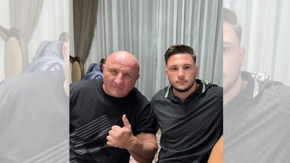 Un bărbat vârstnic cu tricou negru face gestul cu degetul mare în sus lângă un bărbat tânăr cu tricou gri în interiorul unei încăperi.
