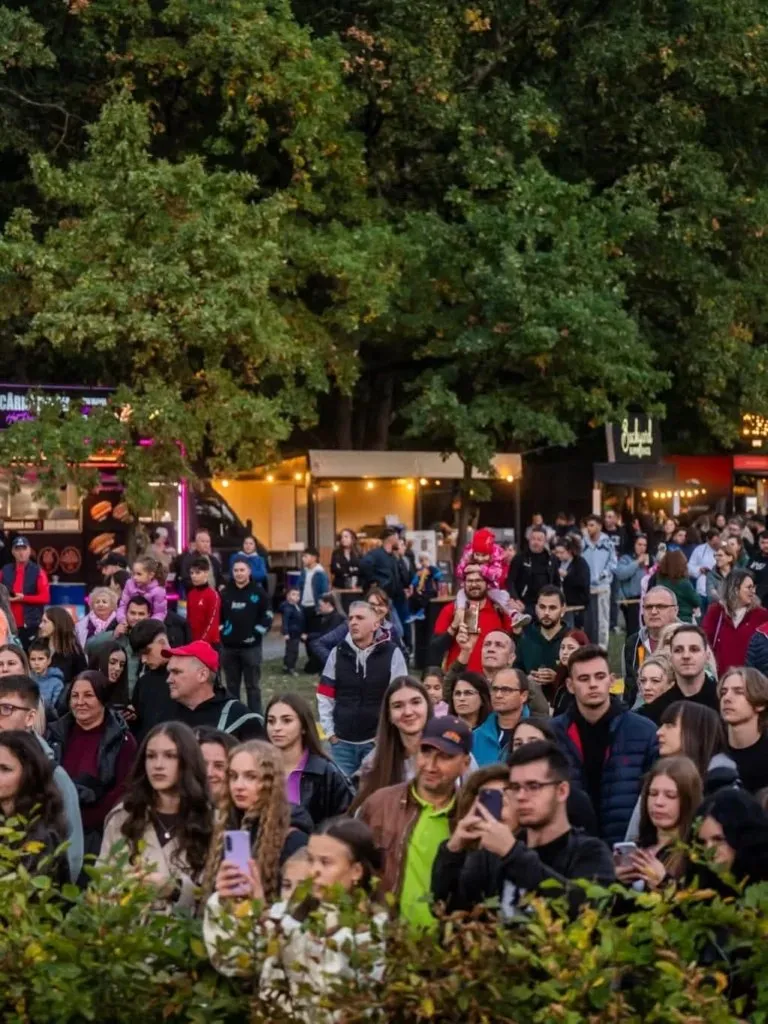 O mulțime de oameni stă în fața unor containere de tip food truck sub copacii înalți într-un spațiu verde.