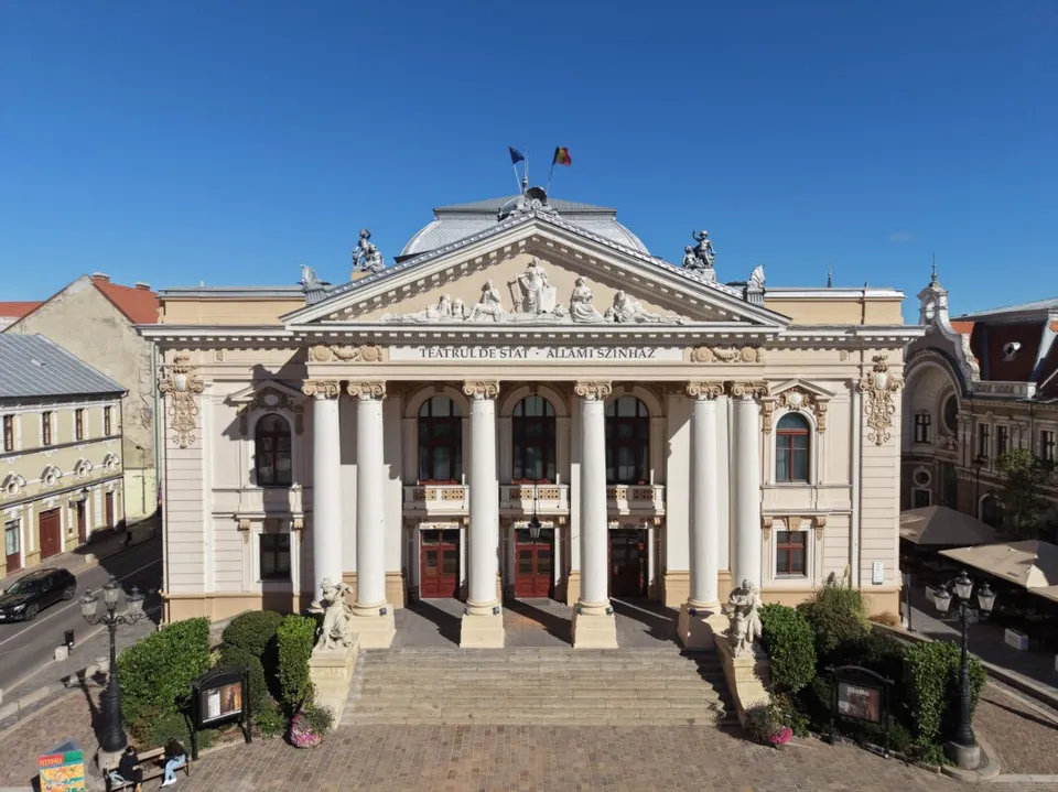 O clădire cu coloane albe, fațadă decorată și un fronton sculptat sub un cer senin.