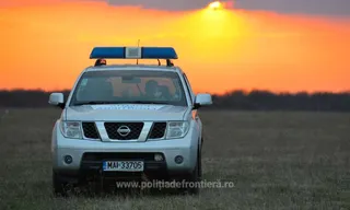 Un vehicul de poliție de culoare argintie cu bară de lumini albastră este parcat pe un câmp sub un cer portocaliu la apus.