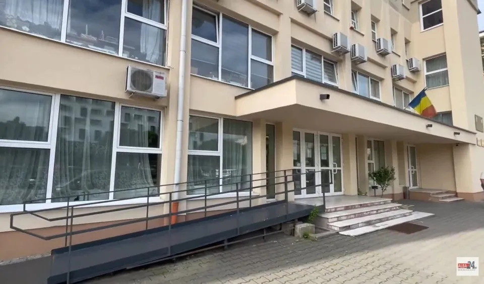 O clădire cu ferestre mari, o rampă de acces metalică și câteva trepte de beton la intrare.