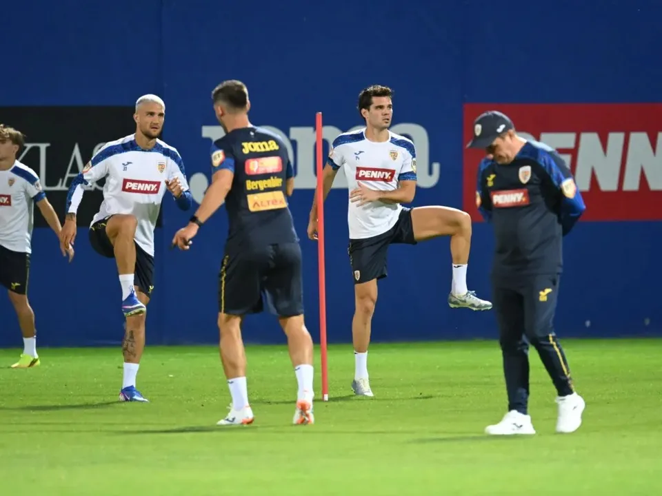 Câțiva bărbați în echipamente sportive albastre și albe execută exerciții de stretching pe un teren de fotbal verde.