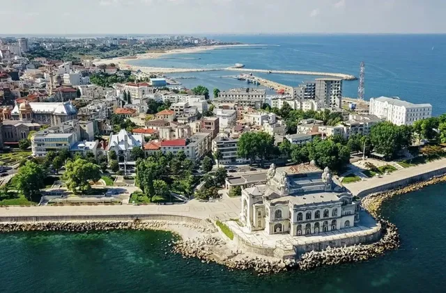Vedere aeriană a unui oraș de coastă cu clădiri diverse, vegetație și un port situat lângă o mare de culoare albastră sub un cer senin.