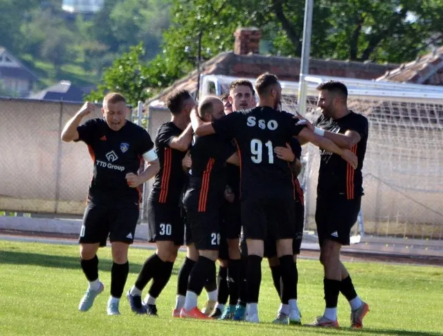 Jucători de fotbal în uniforme negre și portocalii se adună pe gazon în fața porții.