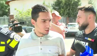 Un bărbat cu tricou alb polo este filmat de un reporter cu microfon și o cameră video în presence a unui polițist.