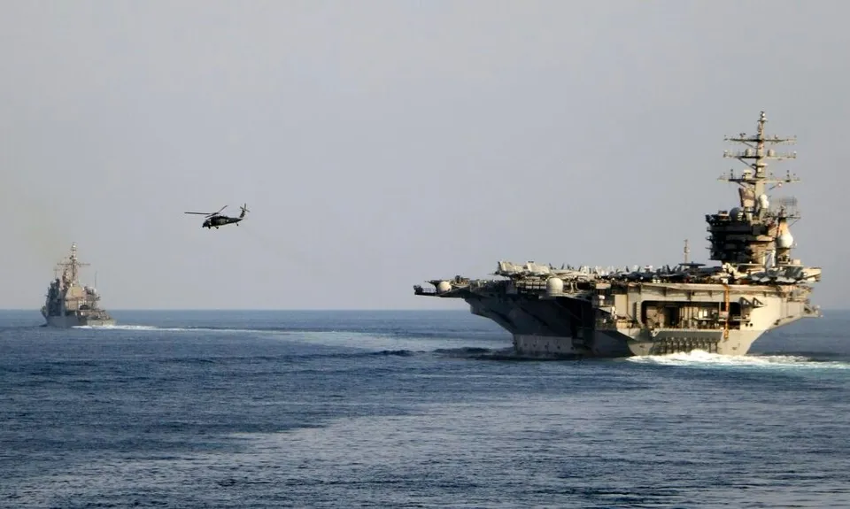 Un portavion mare navighează pe apă lângă un elicopter și un alt vas militar mai mic în depărtare sub un cer senin.