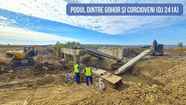 Două persoane purtând veste fluorescente stau lângă o structură de beton și o rampă metalică într-un șantier cu utilaje de construcții.