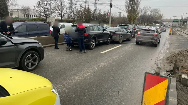 Mai multe autoturisme și o autoutilitară sunt oprite pe un drum asfaltat lângă un trotuar, în prezența unor persoane.