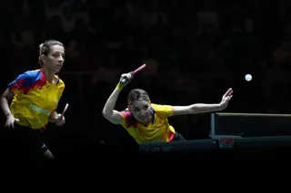 Două femei în echipamente galbene și albastre joacă tenis de masă lângă o masă de joc într-o sală întunecată.