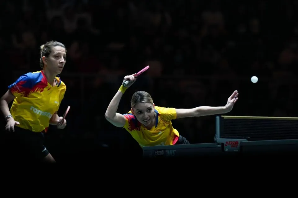 Două femei în echipamente galbene și albastre joacă tenis de masă lângă o masă de joc într-o sală întunecată.