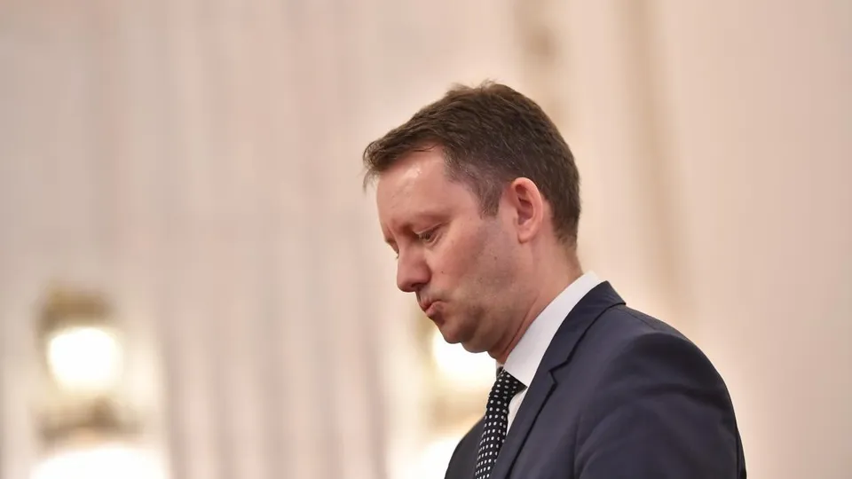 Un bărbat în costum bleumarin și cravată cu puncte, privind în jos cu un expresie serioasă, pe un fundal deschis și neclar.