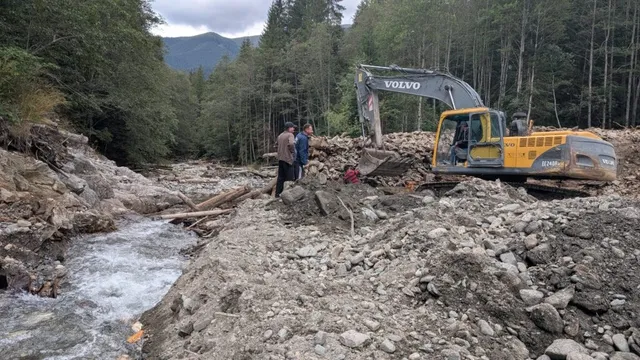 O excavatoare galbenă de marcă Volvo lucrează pe un teren pietros lângă un curs de apă înconjurat de pădure.