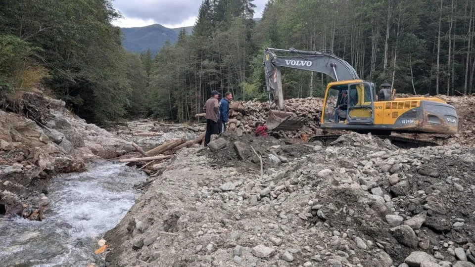 O excavatoare galbenă de marcă Volvo lucrează pe un teren pietros lângă un curs de apă înconjurat de pădure.
