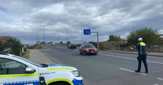 Un polițist cu vestă reflectorizantă stă pe marginea unei drumuri asfaltate lângă un automobil roșu sub un cer înnorat.