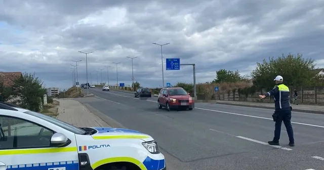 Un polițist cu vestă reflectorizantă stă pe marginea unei drumuri asfaltate lângă un automobil roșu sub un cer înnorat.