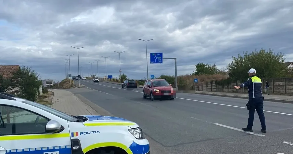 Un polițist cu vestă reflectorizantă stă pe marginea unei drumuri asfaltate lângă un automobil roșu sub un cer înnorat.