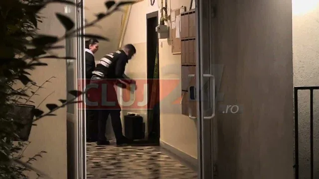 Un bărbat cu o cămașă cu dungi orizontale este văzut în interiorul unui hol, lângă o ușă deschisă, lângă o altă persoană.