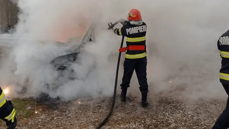 Un pompier cu echipament de protecție și un furtun de stingere acționează asupra unui vehicul acoperit de fum dens.