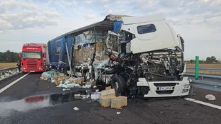 Două camioane sunt implicate într-o coliziune pe o autostradă, având diverse obiecte și cutii împrăștiate pe carosabil lângă vehiculele avariate.