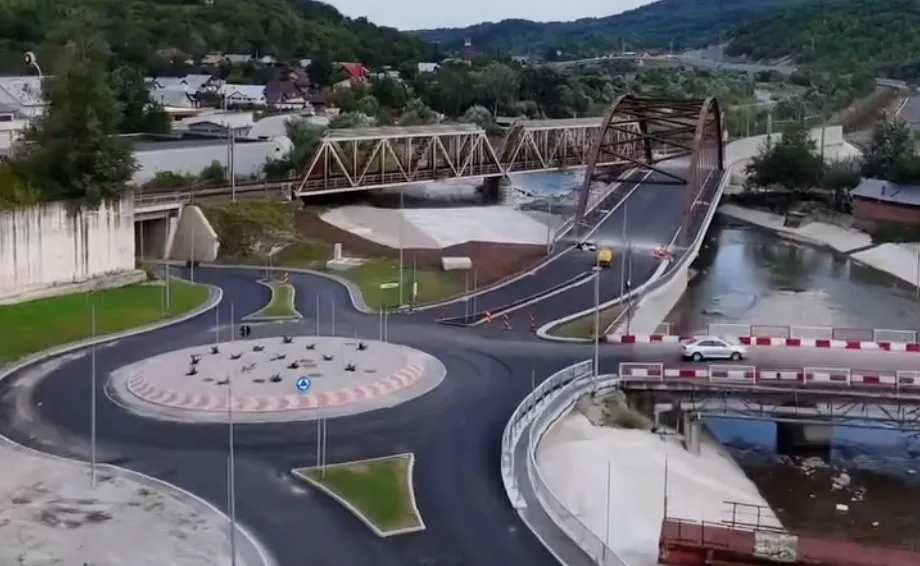 O vedere aeriană prezintă o intersecție cu sens giratoriu, drumuri asfaltate noi, un pod metalic și un râu care traversează un peisaj cu dealuri verzi.