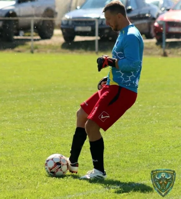 Un fotbalist într-un echipament albastru și roșu stă pe terenul de fotbal lângă un balon aflat pe iarbă.