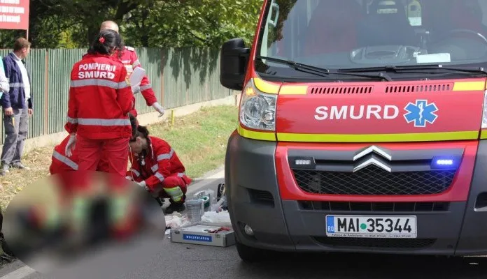 O ambulanță roșie cu inscripția SMURD stă pe marginea drumului lângă personal medical în uniformă roșie care se ocupă de o persoană întinsă pe asfalt.