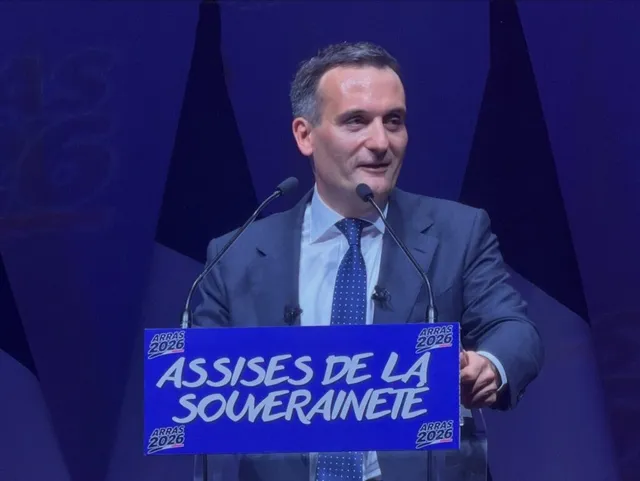 Un bărbat cu părul scurt și costum gri stă în spatele unui podium albastru pe care scrie text alb.