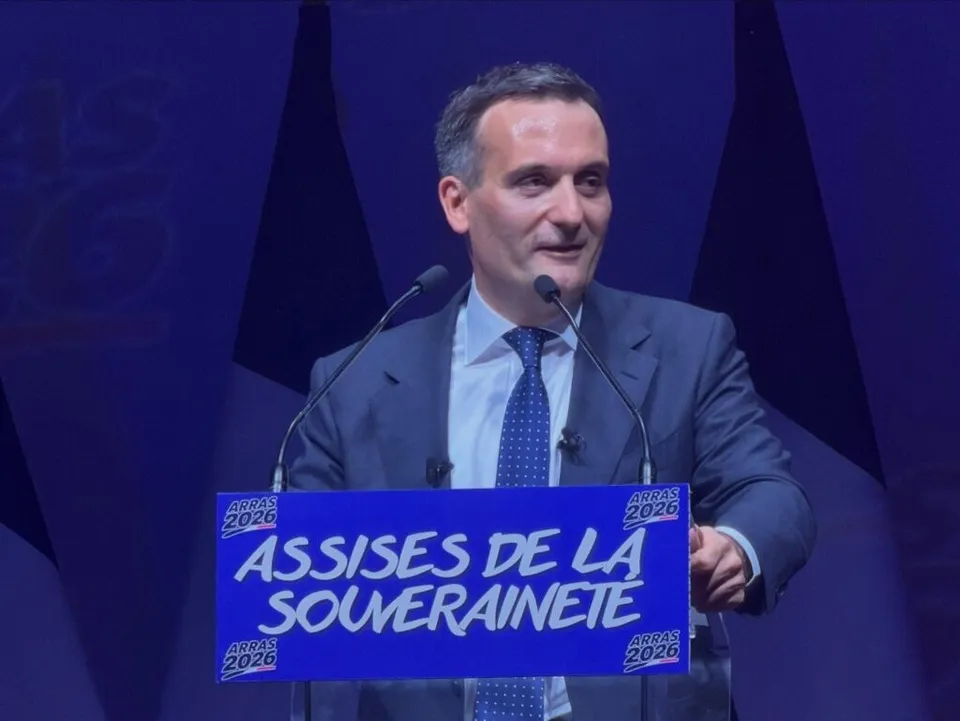 Un bărbat cu părul scurt și costum gri stă în spatele unui podium albastru pe care scrie text alb.