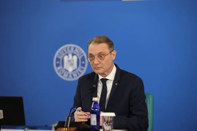 Un bărbat cu ochelari și costum închis stă la o masă în fața unui fundal albastru cu un emblema circulară.
