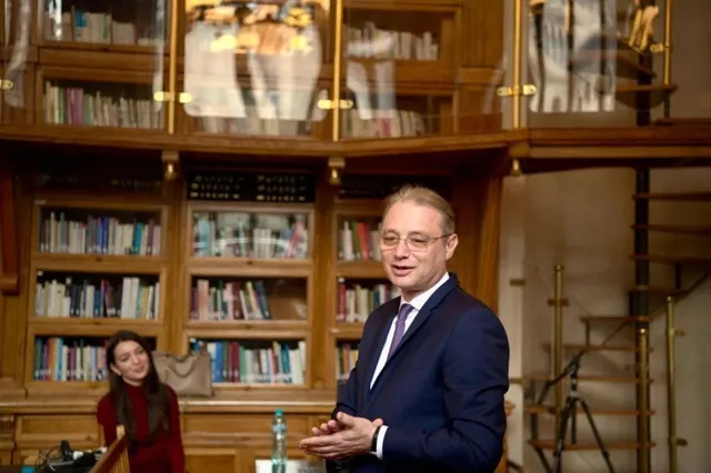 Un bărbat cu ochelari și costum închis la culoare stă în fața unor rafturi de bibliotecă pline cu cărți colorate.